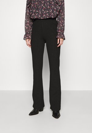 Mujer con pantalones negros de rayas a la cintura alta, blusa floral de manga larga y botines negros de tacón, de pie frente a un fondo liso.