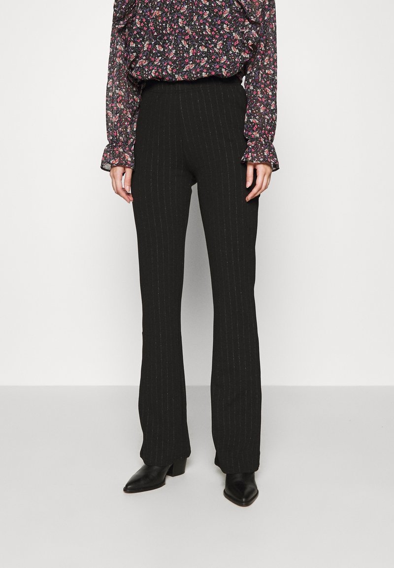 Femme portant un pantalon noir à rayures fines taille haute, un chemisier à manches longues fleuri et des bottines noires à talons, debout devant un fond uni.
