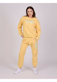 Gul sweatshirt og matchende sweatpants. Blød tekstur med afslappet pasform. Blå trykt tekst foran og på siden. Hvide sneakers fuldender looket.