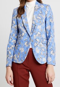Light blue floral blazer with a structured design, featuring gold accents, a single button, and two front pockets. Worn over a white turtleneck.
