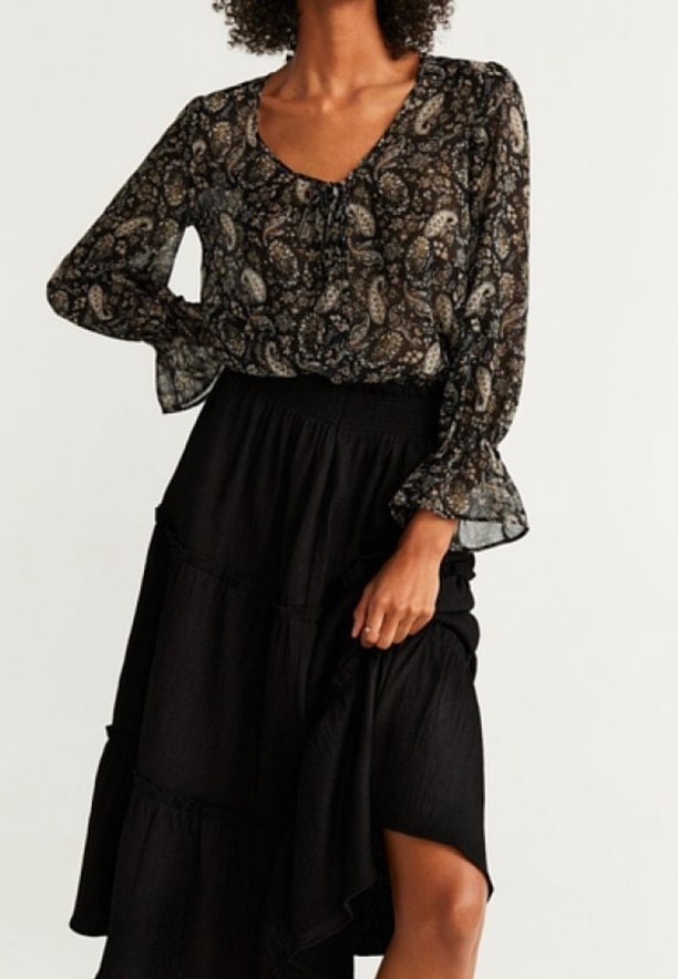 Femme portant un chemisier transparent à manches longues à motif paisley, rentré dans une jupe noire à volants, tenant la jupe d'une main.