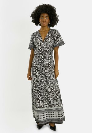 Femme avec coiffure afro portant une robe maxi à motifs noirs et blancs à manches courtes et des chaussures noires pointues, debout sur fond blanc.