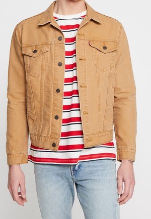 Personne portant une veste en jean beige boutonnée sur un t-shirt rayé rouge, blanc et noir et un jean bleu clair, debout devant un fond uni.