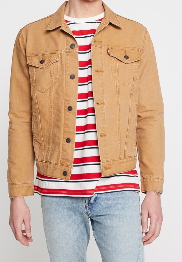 Personne portant une veste en jean beige boutonnée sur un t-shirt rayé rouge, blanc et noir et un jean bleu clair, debout devant un fond uni.