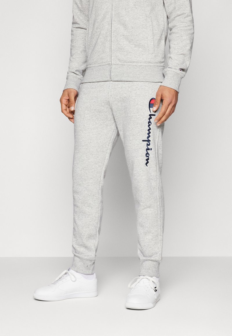 Grå joggebukser laget av mykt stoff, med en brodert "Champion"-logo i marineblått og rødt på siden, og ribbede ankelmansjetter.