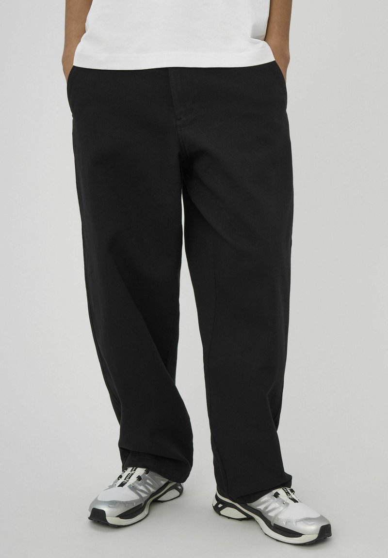 Pantalon noir à coupe ample en tissu mélangé coton avec poches latérales, associé à des chaussures de sport argentées et noires, posé sur un fond neutre.