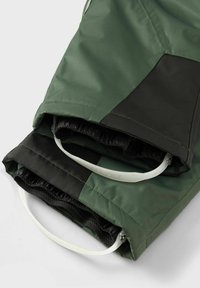 Žalia vandeniui atspari striukė su tekstūruotu juodu elementu, elastingomis rankogaliais ir raištelių detalėmis apačioje, leidžiančiomis reguliuoti dydį.