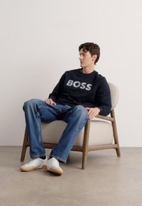 Jūras spēku svārki ar uzrakstu "BOSS" gaiši pelēkā krāsā, zilas džinsus un baltas kedas. Sēž uz gaiši pelēkas tapecētas krēsliem ar koka kājām.