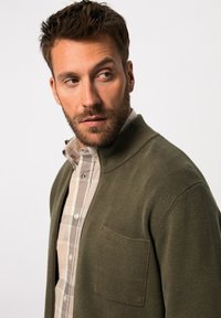 Cardigan verde oliva de punto texturizado, con cierre de cremallera en la parte delantera y un bolsillo en el pecho, superpuesto a una camisa de cuadros en tonos neutros.