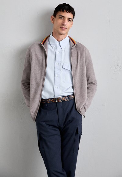Cárdigan de punto gris claro con cremallera y cuello a rayas multicolores, llevado sobre una camisa de botones azul claro, combinado con pantalones azul marino y un cinturón trenzado.