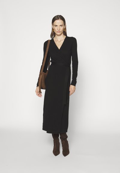 Robe noire à manches longues et à ceinture à la taille. Le mannequin porte des bottines en daim marron et un sac à bandoulière marron.