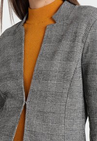 Blazer gris de cuadros con un corte estructurado, que presenta un sutil patrón de cuadros y un cuello afilado. Combinado con un cuello alto mostaza de canalé debajo.