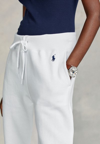 Άτομο που φοράει λευκό παντελόνι φόρμας με κορδόνι και μπλε σήμα polo, και ρολόι με καφέ δερμάτινο λουράκι στον δεξί του καρπό, έχοντας το χέρι στην τσέπη.