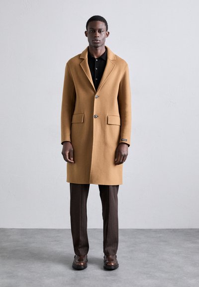 Manteau en mélange de laine marron avec un col cranté, deux poches avant et fermeture à boutons, associé à un pantalon à rayures noires et des chaussures.