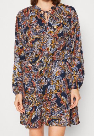 Woman wearing long-sleeve knee-length dress with blue, orange, yellow, and white abstract floral pattern and tie neckline.