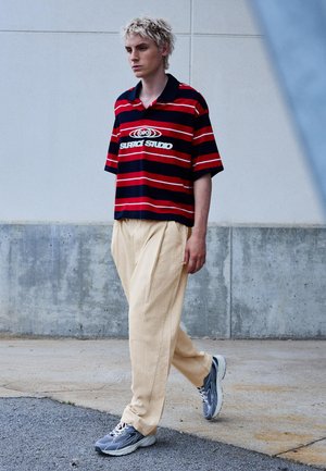 Mladý muž kráča popri betónovej stene, má na sebe červené a námornícky modré pruhované tričko s textom, béžové nohavice a sivé tenisky.