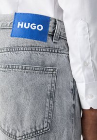 Grå denimjeans har en blå etikett med "HUGO" på bakliv, og er kombinert med en hvit skjorte med knapper.