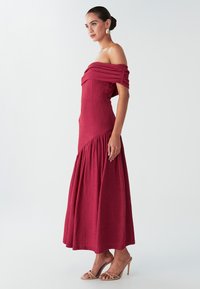 Schulterfreies, erdbeerrotes Kleid aus strukturiertem Stoff; tailliertes Oberteil geht in einen ausgestellten Rock über. Getragen mit nude-farbenen Riemchen-Heels.