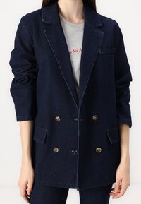 Veste blazer croisée en denim foncé avec un col à revers cranté, deux poches avant et des boutons dorés. Portée sur un t-shirt gris clair.