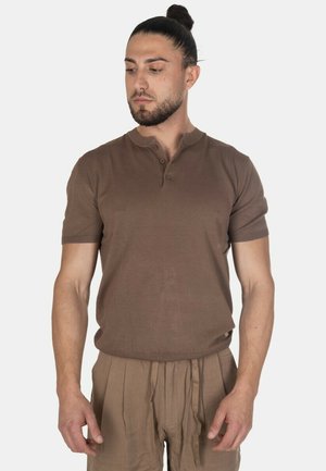 Uomo con chignon che indossa una maglietta henley marrone a maniche corte e pantaloni plissettati marroni abbinati, guarda in basso verso sinistra.