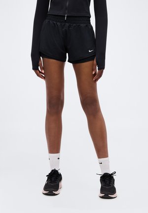 Osoba ubrana w czarne sportowe spodenki, czarną bluzę rozpinaną, białe skarpety Nike i czarne buty do biegania stojąca na jednolitym tle.