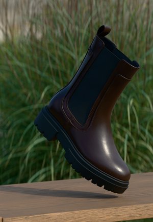 Botte Chelsea en cuir marron foncé avec une semelle noire épaisse sur une surface en bois, arrière-plan flou avec de l'herbe haute verte.
