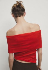 Haut rouge décolleté en tissu doux, présentant un design froncé. L'arrière-plan est uni et clair.