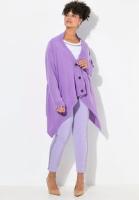 Cardigan viola con collare a scialle, tre bottoni e orlo asimmetrico, abbinato a pantaloni lilla e décolleté color nude. Tessuto morbido.