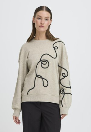 Vrouw die een beige sweatshirt met abstract zwart lijnpatroon en een zwarte broek draagt, staand tegen een effen witte achtergrond.
