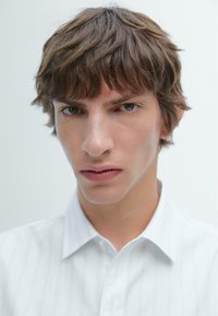 Weißes Hemd mit Nadelstreifen und Kragen, aus strukturiertem Stoff und in taillierter Passform. Nahaufnahme einer Person mit kurzen, zerzausten braunen Haaren.