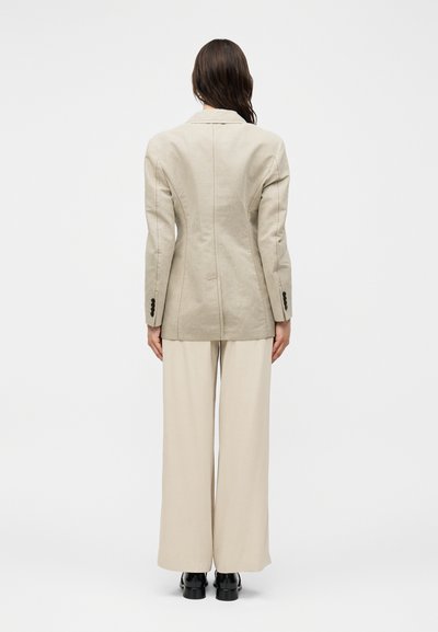 Γυναίκα φορώντας μπεζ ραμμένο σακάκι και φαρδιά παντελόνια, στέκεται με την πλάτη προς τον θεατή σε λευκό φόντο, εμφανείς μαύρες παπούτσια.