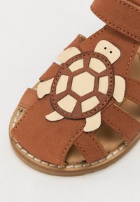 Brune lærsandaler med en skildpadde-design på remmen, med udskårne detaljer og en tekstureret overflade. Justerbar velcrolukning.
