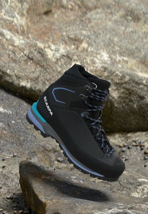 Bota de senderismo Scarpa negra y azul con cordones estampados suspendida sobre terreno rocoso y fondo de piedra.