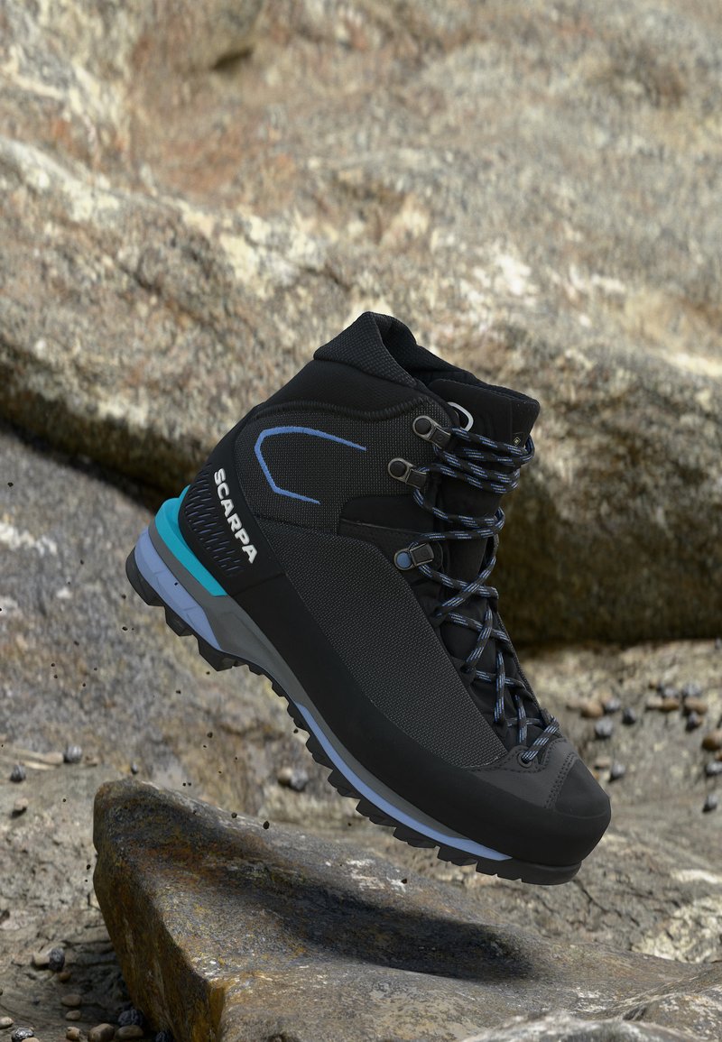 Μαύρο και μπλε μποτάκι πεζοπορίας Scarpa με κορδόνια με σχέδια που κρέμεται πάνω από βραχώδες έδαφος και πέτρινο φόντο.