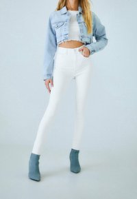 Veste en denim bleu clair raccourcie, jean skinny taille haute blanc, et bottines grises. Lignes épurées avec des détails d'ourlet effiloché sur la veste.