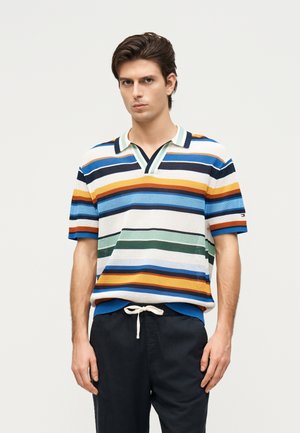 Joven que lleva un polo de punto a rayas de manga corta con colores azul, verde, naranja y blanco, combinado con pantalones negros con cordón.