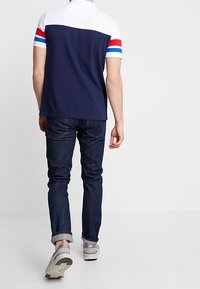 Navy blue polo shirt with white upper section and red, blue striped sleeve accents, paired with dark denim jeans and grey sneakers.