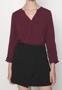 Blouse texturée bordeaux avec un col en V et des manches trois-quarts, associée à une jupe portefeuille noire avec des poches avant et une silhouette épurée.