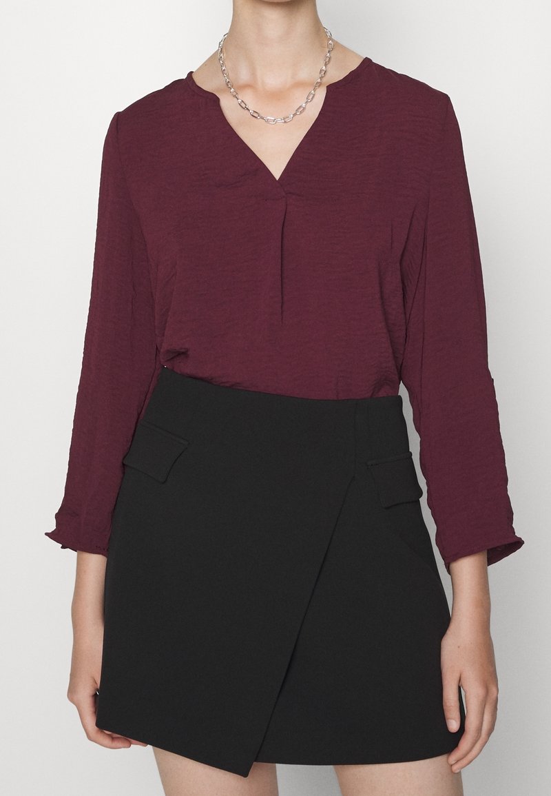 Blouse texturée bordeaux avec un col en V et des manches trois-quarts, associée à une jupe portefeuille noire avec des poches avant et une silhouette épurée.