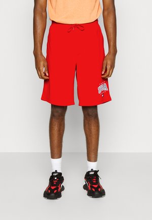 Pantaloni sportivi rossi con vita elasticizzata e lacci, con una grafica USA in bianco e blu. Indossati con scarpe da ginnastica nere e rosse.