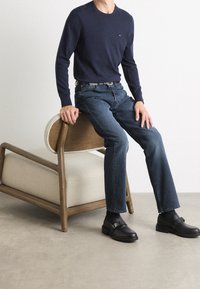 Pull marine, jean bleu foncé et chaussures noires avec une boucle, assis sur une chaise en bois avec un coussin en tissu clair. Design simple et moderne.