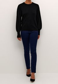 Black long-sleeve sweater with ribbed cuffs, paired with fitted dark blue jeans and black high-heel shoes, model standing against a neutral background.
