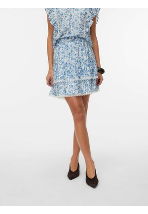 Femme portant une robe courte bleue à motifs floraux avec finition en dentelle et des talons noirs à bout pointu, debout les jambes croisées.