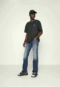 Νέος άνδρας φορώντας μαύρο καπέλο, υπερμεγέθες μαύρο t-shirt, μπλε τζιν και μαύρα σανδάλι με λευκές κάλτσες, στέκεται μπροστά σε απλό φωτεινό φόντο.