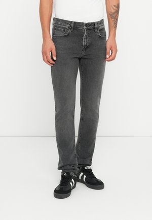 Pantalones vaqueros de mezclilla gris con corte ajustado, cinco bolsillos y un ligero desvanecimiento. Llevados con zapatillas negras con rayas blancas.