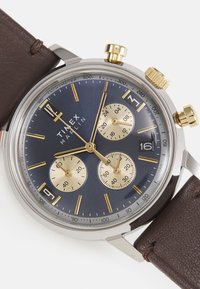 Montre chronographe avec un cadran bleu marine, des accents dorés, trois sous-cadrans et un bracelet en cuir marron. Boîtier en acier inoxydable avec tachymètre.