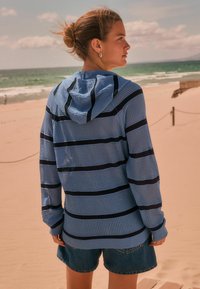  Lichtblauwe, gestreepte hoodie van een textuurstof. Voorzien van een capuchon en lange mouwen. Gepaard met denim shorts. Strandachtergrond zichtbaar.