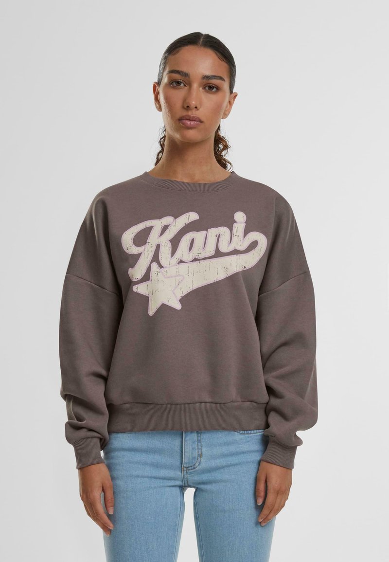 Maglione grigio con un grande logo retro "Kani" bianco e una grafica a stella, con polsini e orlo a costine. Abbinato a jeans azzurri chiari.