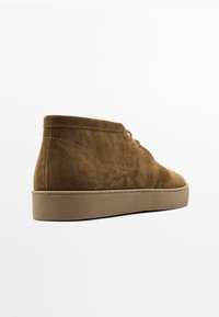 Bottines en suède marron avec une finition texturée, bout arrondi, fermeture à lacets à l'avant, et une semelle en caoutchouc beige, présentant des détails de couture minimalistes.