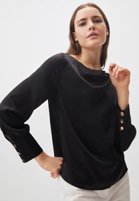 Blusa nera con scollatura drappeggiata, maniche lunghe e bottoni dorati sui polsini. Tessuto morbido, vestibilità comoda e orlo curvo.